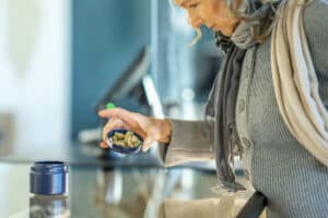 A mature woman leans in over a counter and reaches for some cannabis buds that have been laid out by the associate in the product lid. The woman is inspecting the product before purchasing from the legal retailer.