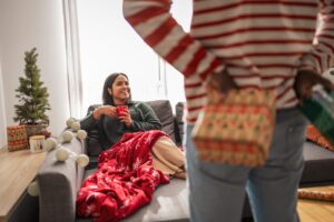 A woman in a cozy living room receives a surprise gift, wrapped in holiday paper, while enjoying a warm beverage. The scene is filled with festive decorations and Christmas spirit.